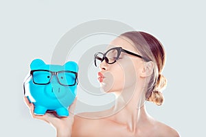 Glasses sale concept. Happy woman kissing piggy bank wearing eye wear glasses. Female model  on light blue background