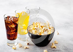 Glasses of soda drink with ice cubes and black bowl of popcorn snack on stone kitchen table background. Cola and orange and clear