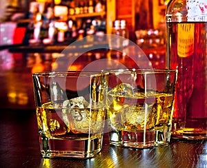 Glasses of whiskey with ice on bar table near whiskey bottle on warm atmosphere