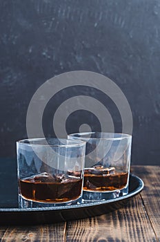 Glasses of whiskey with ice cubes on a black tray and wooden table/Two Glasses of whiskey with ice cubes on a black tray and