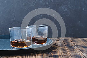 Glasses of whiskey with ice cubes on a black tray and wooden table/Two Glasses of whiskey with ice cubes on a black tray and