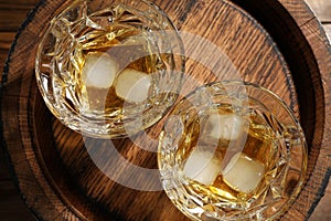Glasses of whiskey with ice cubes on wooden barrel, top view