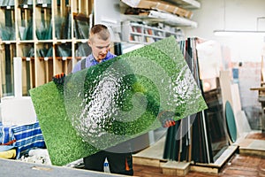 Glazier worker holding a green glass pane in workshop.