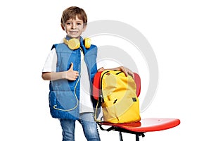 Gleeful little boy with headphone and backpack stay near red chair on white background. Back to School. Show thumb up.
