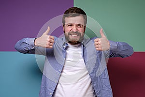 Gloating Caucasian man makes an insincere approving gesture with hands, raises thumbs up and grins teeth, squinting eyes