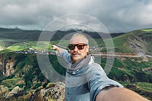 Global Travel Concept. Young Traveler Man With A Beard And Sunglasses Take A Selfie On A Background Of A Mountain Landscape