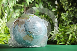 Globe in plastic bag on table against green leaves, space for text. Environmental conservation