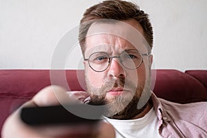 Gloomy man in glasses holds a TV remote control and switches channels displeasedly. Close-up.