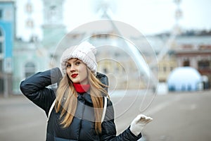 Glorious blonde model with red lipstick wearing knitted cap, enjoying city walk. Empty space