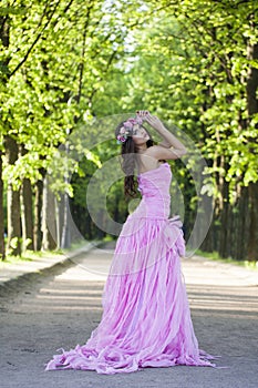 Glorious female model with bright makeup, curly hair and pink flower wreath on green summer foliage background. Healthy woman in