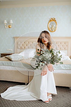 Glorious lady in long wedding gown decorated with feathers sitting on edge of double bed with bouquet of white flowers.