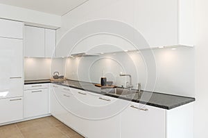 Glossy white minimalist modern kitchen with a black marble countertop. Spotlights illuminate the countertop, above which are two