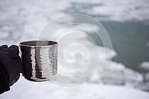 Gloved hand holding warm beverage with icy water in distance