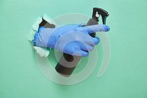 A gloved hand holds a disinfectant in a black spray bottle. Green paper background with torn hole