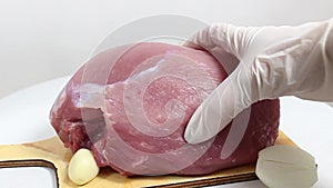 A gloved hand presses on a raw piece of pork that is lying on a garlic chopping board.