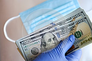 Gloved hands hold hundred-dollar bills against the background of a medical mask.