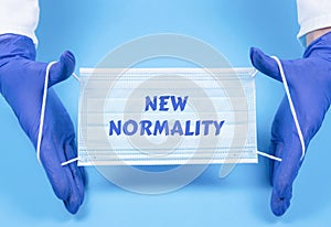 Gloved hands holding medical mask on blue background. Text design New normality, lettering on protective mask for post covid-19