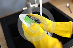 Gloved hands wash dishes under tap with sponge