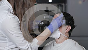 A gloved makeup artist corrects, shapes, and colors the eyebrows of the client in a protective mask at home during the