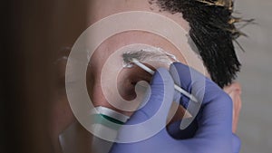 A gloved makeup artist corrects, shapes, and colors the eyebrows of the client in a protective mask at home during the