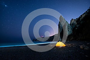 Glowing camping tent on a beautiful sea shore with rocks at night under a starry sky