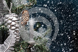 Glowing candle on Christmas tree with pine cone by night on background of falling snow.