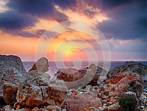 a glowing golden sunset surrounded by illuminated clouds over a calm Mediterranean sea with a foreground of coastal rocks colored