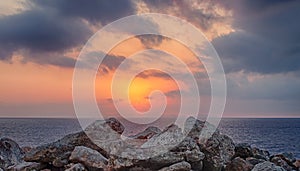 a glowing golden sunset surrounded by illuminated clouds over a calm Mediterranean sea with a foreground of coastal rocks colored