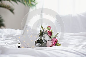 Glowing LED star and a bouquet of artificial peonies on a white bed in the interior