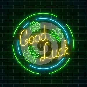 Glowing neon sign with geed luck wish and four-leaf clovers in circle frames on dark brick wall background.