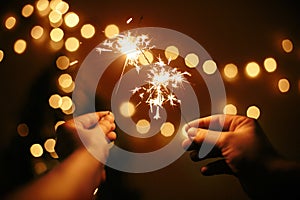 Glowing sparklers in hands on background of golden christmas tree lights, couple celebrating in dark festive room. Happy New Year