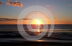 Sunset with lens flare over a calm blue sea and beach with illuminated orange clouds