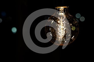 Glowing vintage old one tungsten lightbulb hanged on ceiling against black wall bokeh background. Retro styled decoration electric