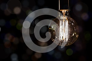 Glowing vintage old one tungsten lightbulb hanged on ceiling against black wall bokeh background. Retro styled decoration electric