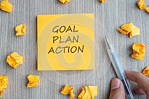 Goal, Plan, Action word on yellow note with Businessman holding pen and crumbled paper on wooden table background. Business Vision