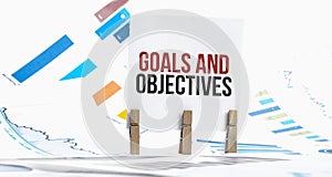 Goals and Objectives text on paper sheet with chart, dice, spectacles, pen, laptop and blue and yellow push pin on wooden table -