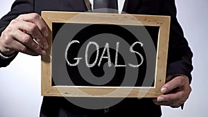 Goals written on blackboard, man in classic suit holding sign, business concept