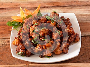 Gobi Manchurian, Popular indo-chinese food made of cauliflower florets, served in a white plate over a rustic wooden table.