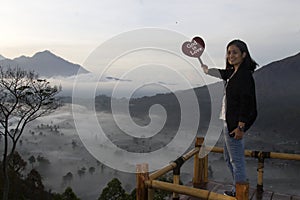 God is Love. Happy woman standing with red love heart sign in the mountains at sunrise on a misty morning. photo