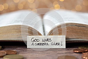 God loves a cheerful giver, handwritten text note in front of open holy bible book and golden coin money with bokeh background