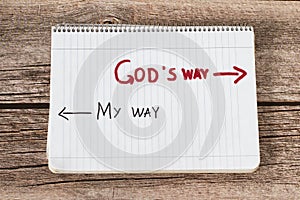 God`s way, a handwritten text quote and arrows in a white notebook page placed on a wooden background