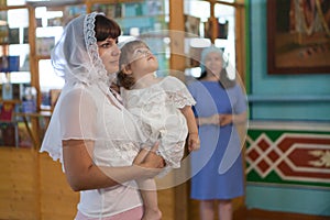 Godmother with goddaughter stand in the Orthodox church. Mother standing on the side. Ceremony of sacrament of initiation into the