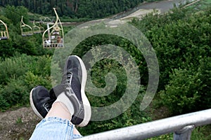 Going down from mountain top on cableway. Person legs and cable car seat, funicular over green mountain valley, top view. Active