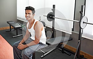 This is going to be an awesome workout. A young ethnic man sitting on a weight bench at a gym.