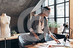 This is going to be a long day. Attractive young woman in eyewear using laptop while standing near the desk in her workshop