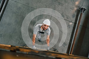 Going up on the ladder, top view. Young factory worker in grey uniform