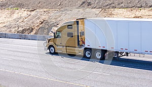 Gold big rig modern semi truck transporting cargo in semi trailer driving on the multiline highway with developed hill land