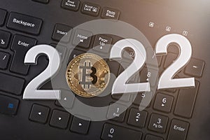 A gold bitcoin is placed on a dark-colored keyboard. The year 2022 is laid out in metal numbers. close-up