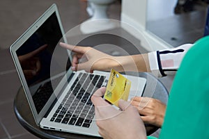 Gold credit card in the hand of a woman in a green dress. Her friend pointing finger at laptop computer screen in the background