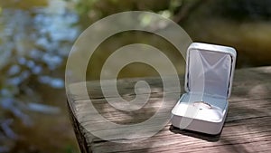 Gold engagement ring in white box sits on wooden table by river